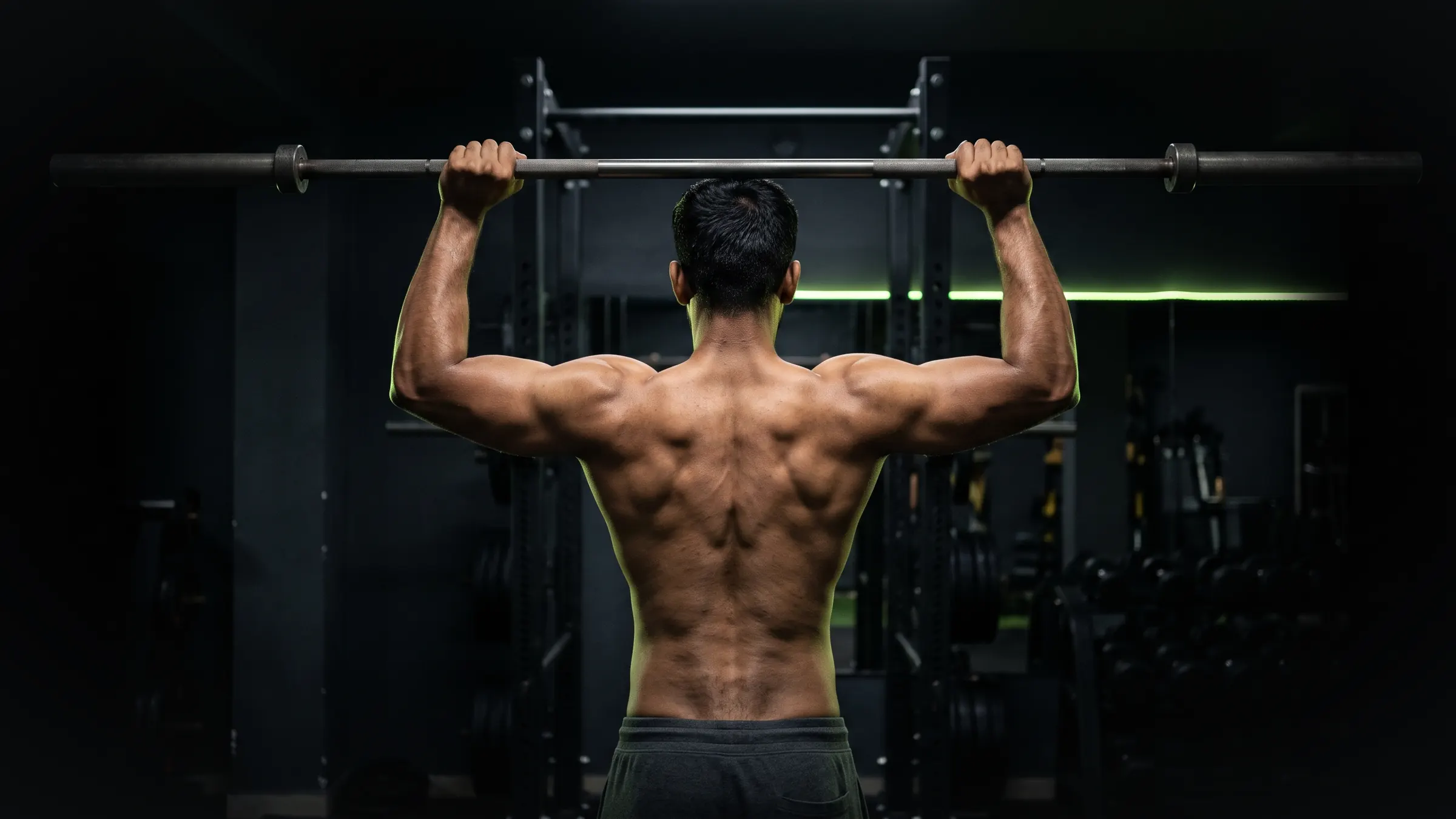 Athlete seen from behind lifting a barbell overhead in a dark gym